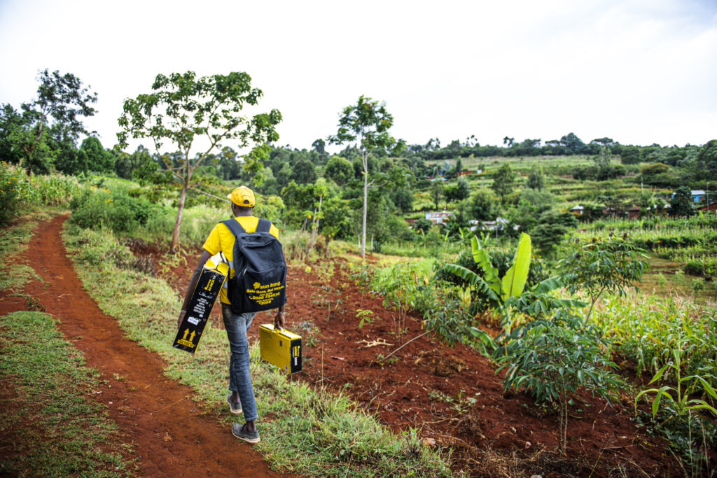 A Sun King field agents walks down a dirt road in Kenya carrying Sun King products surrounded by verdant nature and trees.