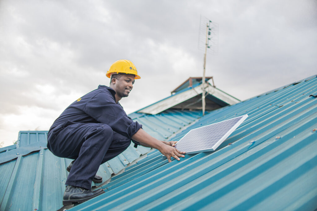 A Sun King engineer in a yellow helmet installs a solar panel on a blue roof in Kenya.