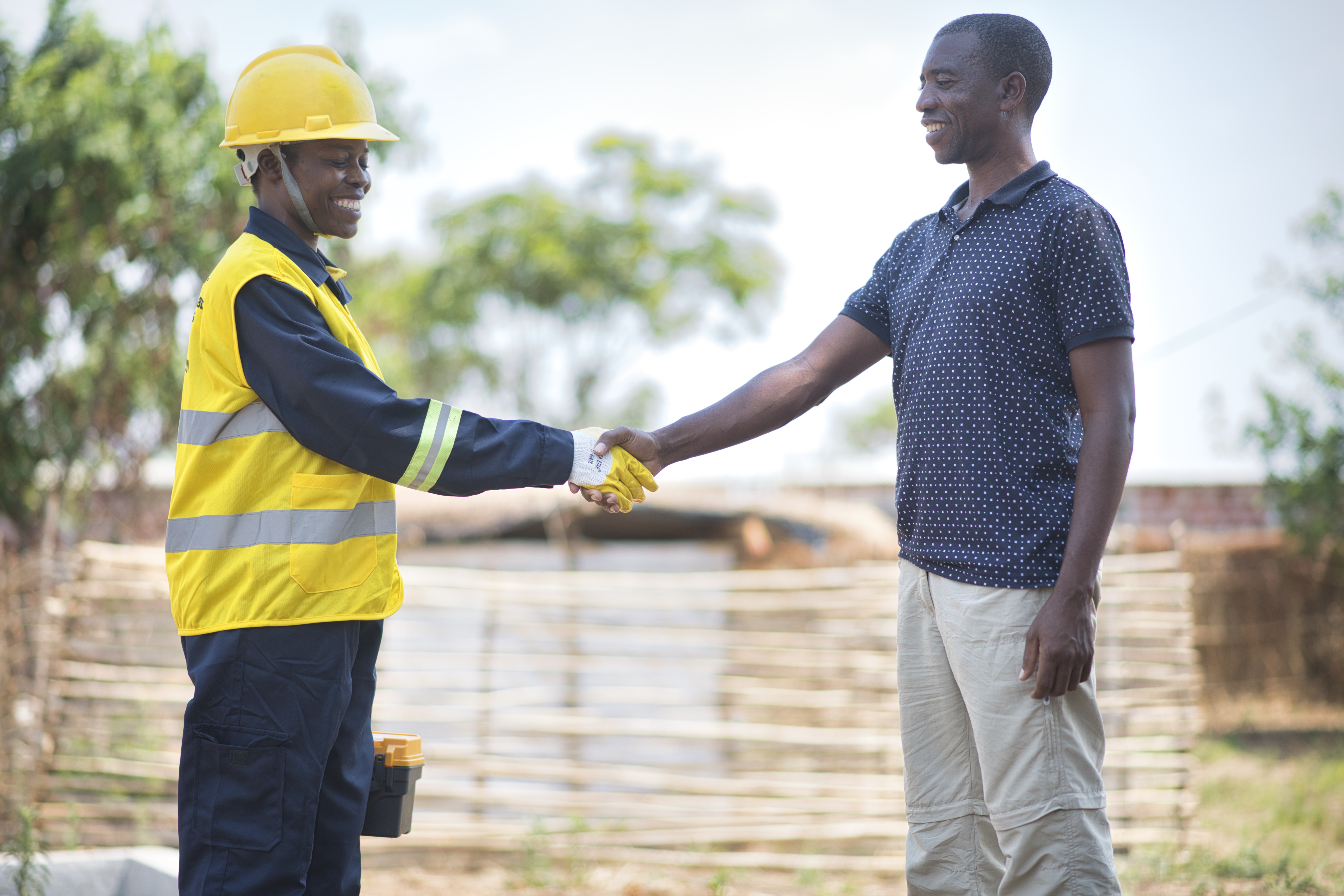 A solar inverter installer in a yellow backet shakes hand with a man in a blue shirt in a rural setting with trees in the background.