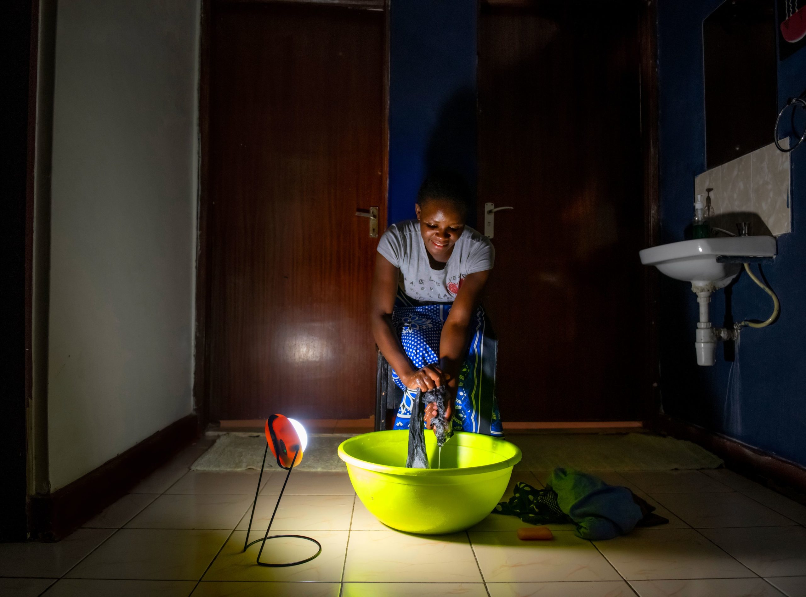 A woman washes her clothes in a lime green basin, using the Sun King Pro 400 solar lantern to light her as she works.
